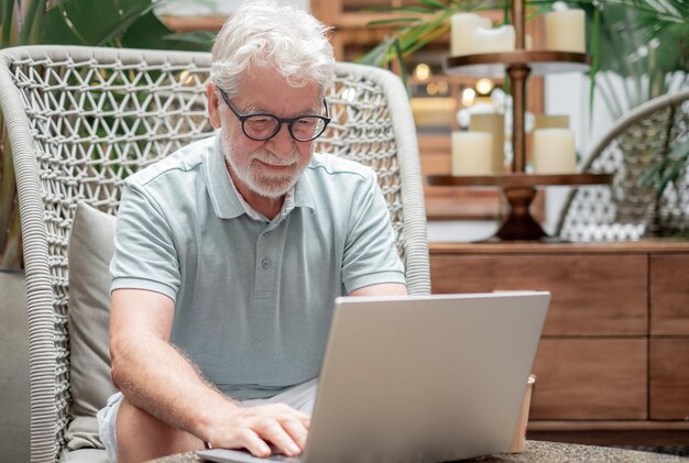 Hombre barbudo mayor sentado en el interior usando una computadora portátil escribiendo en el teclado mientras navega por la red