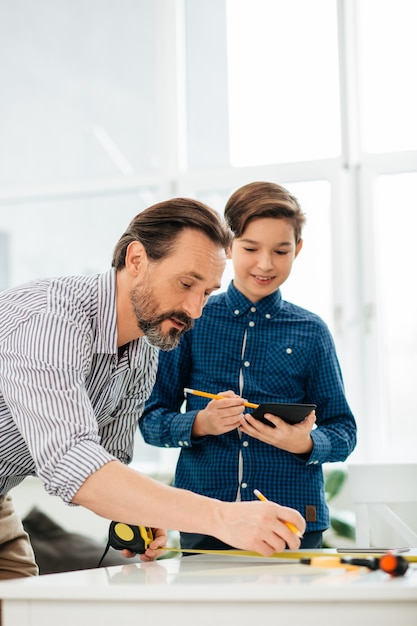 Hombre barbudo de mediana edad entreteniendo a su hijo adolescente sonriente mientras usa una cinta métrica y hace dibujos en la mesa