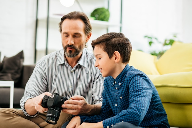 Hombre barbudo de mediana edad sosteniendo una cámara moderna y mostrando los ajustes principales a su curioso hijo interesado