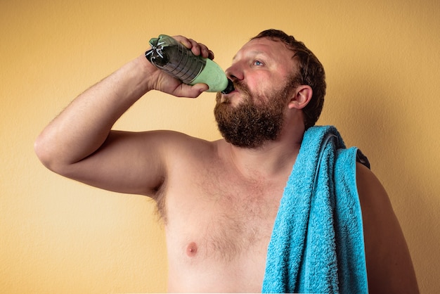 Hombre barbudo medio desnudo cansado y agotado sosteniendo una toalla azul sobre su hombro y beber agua