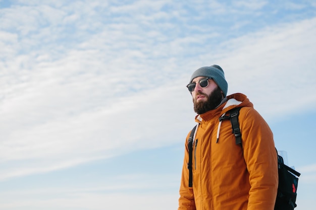 Un hombre barbudo con una mochila con gafas de sol y un gorro cálido contra el fondo del cielo azul El concepto de senderismo y estilo de vida activo