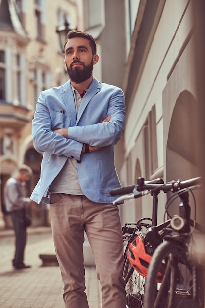 Hombre barbudo de moda con ropa elegante, se para con una bicicleta en una calle antigua en Europa.