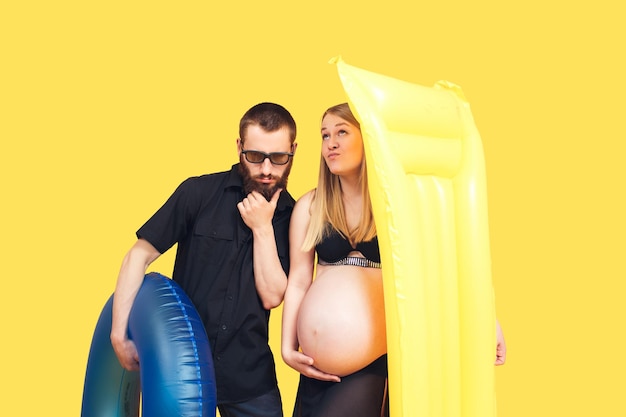 Hombre barbudo y mujer embarazada sosteniendo anillo inflable y colchón sobre fondo amarillo