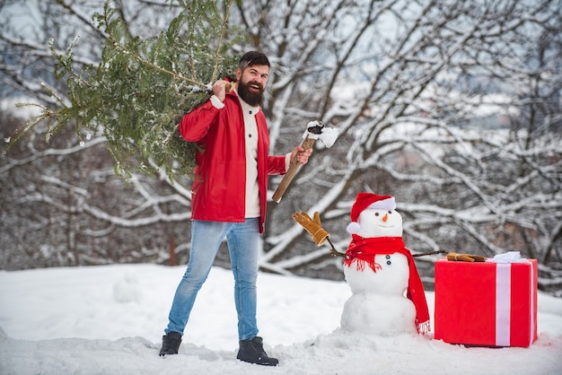 Hombre barbudo con muñeco de nieve lleva árbol de Navidad en el bosque. Un apuesto joven con muñeco de nieve.