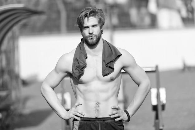 Hombre barbudo con músculos del torso en forma en el estadio Deportista con camiseta azul en el cuello Atleta con vientre y pecho sexy en el soleado al aire libre Entrenamiento y entrenamiento Deporte o fitness y cuidado corporal