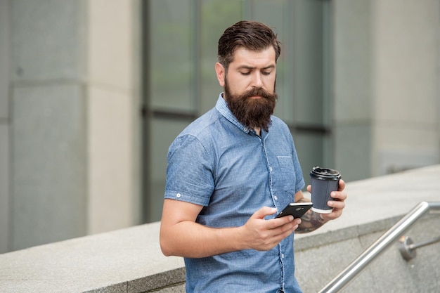 Hombre barbudo navegando en las redes sociales en un dispositivo de teléfono móvil usando Internet móvil mientras bebe café