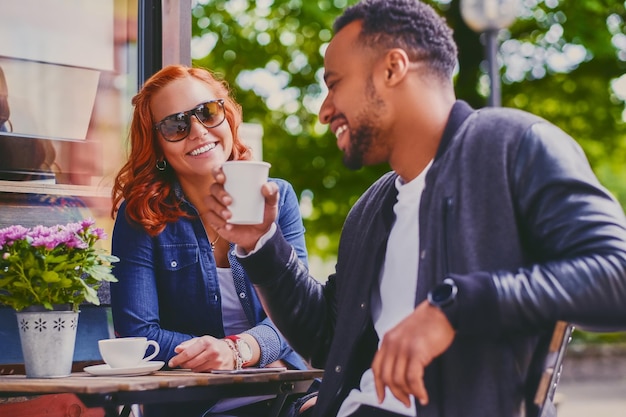 Hombre barbudo negro y mujer pelirroja beben café en una cafetería en una calle.