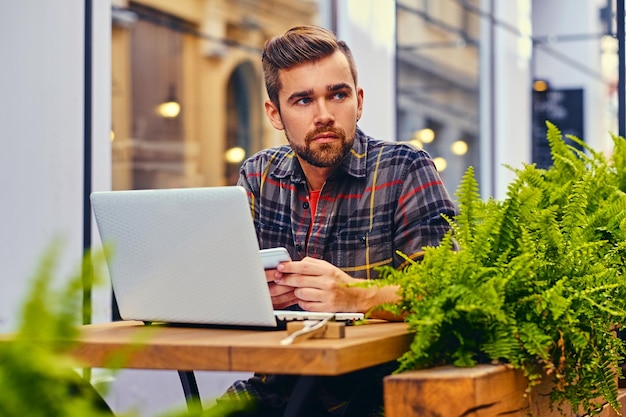 Hombre barbudo de ojos azules que usa una computadora portátil en un café en una calle.