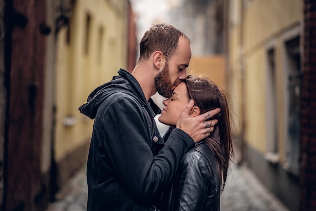 Hombre barbudo positivo besando a una linda morena en una calle de un casco antiguo.