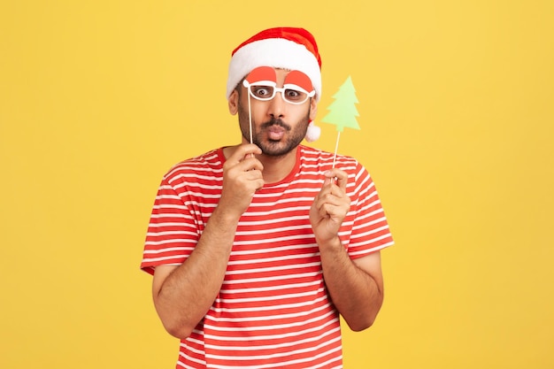 Hombre barbudo positivo en camiseta roja y sombrero de santa claus engañando a las caras que se divierten probándose máscaras de fiesta de navidad en un palo Foto de estudio interior aislada sobre fondo amarillo