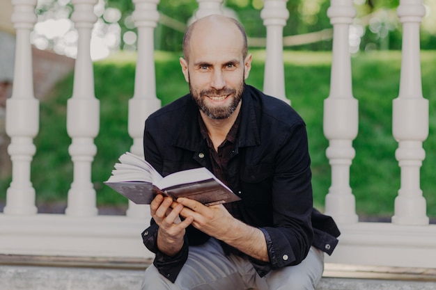 El hombre barbudo positivo mira directamente a la cámara lee un libro muy emocionante e interesante sentado en el fondo del balcón pasa tiempo libre en el jardín de verano Educación y felicidad de la gente