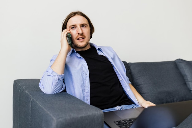 Hombre barbudo positivo con portátil hablando por teléfono inteligente sentado en el sofá en la sala de estar