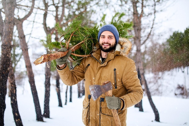 Hombre barbudo que lleva un árbol de navidad recién cortado en el bosque leñador sostiene un hacha y un abeto en