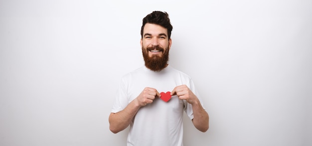 Hombre barbudo que lleva a cabo un pequeño corazón de papel rojo en la camiseta blanca en la pared blanca que sonríe en la cámara.