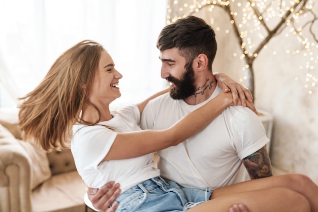 Hombre barbudo romántico que lleva a la mujer en las manos y sonriendo en una habitación luminosa en casa