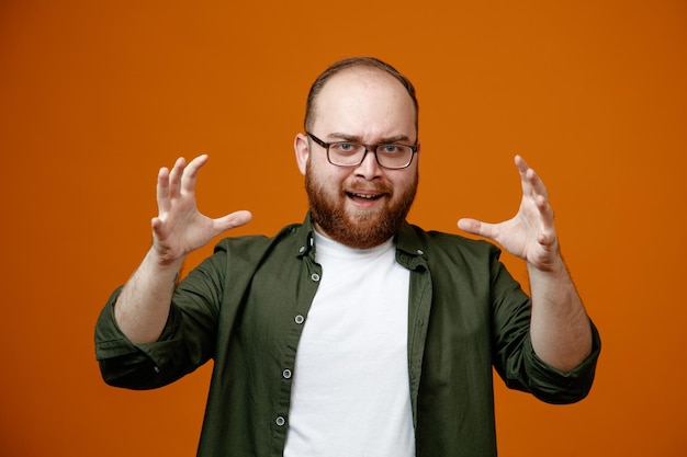 Hombre barbudo en ropa casual con gafas mirando a la cámara aterrador levantando los brazos de pie sobre fondo naranja