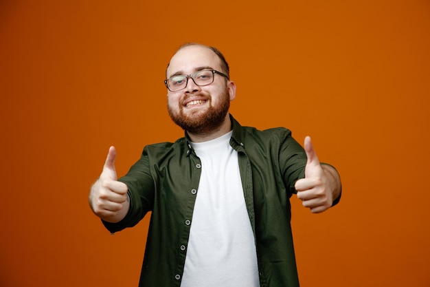 Hombre barbudo en ropa casual con gafas mirando a la cámara sonriendo ampliamente feliz y positivo mostrando los pulgares hacia arriba de pie sobre fondo naranja