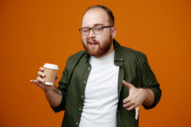 Hombre barbudo en ropa casual con gafas sosteniendo una taza de café mirando a la cámara feliz y fresco sonriendo alegremente de pie sobre fondo naranja