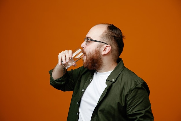 Hombre barbudo en ropa casual con gafas sosteniendo un vaso de agua bebiendo mirando confiado de pie sobre fondo naranja