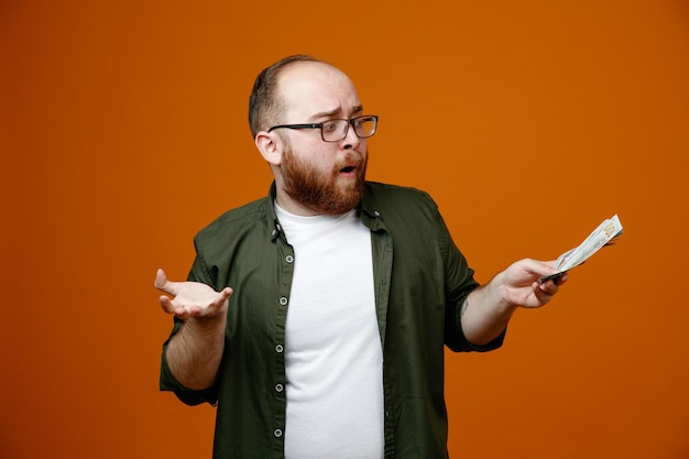 Hombre barbudo con ropa informal y gafas con dinero en efectivo que parece confundido sin respuesta sobre qué hacer de pie sobre un fondo naranja