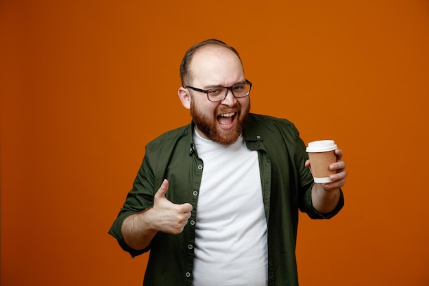 Hombre barbudo con ropa informal con gafas sosteniendo una taza de café mirando a la cámara sonriendo feliz y positivamente mostrando el pulgar hacia arriba de pie sobre un fondo naranja