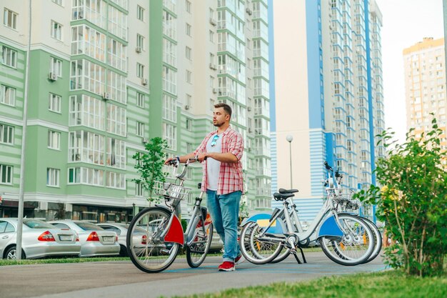 El hombre barbudo con ropa informal usa el transporte ecológico en las calles de la ciudad camina con una bicicleta alquilada en el contexto de un patio con edificios de apartamentos El turista tomó una bicicleta para compartir