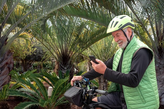 Hombre barbudo senior activo con casco montando su bicicleta en un parque tropical mirando el teléfono móvil