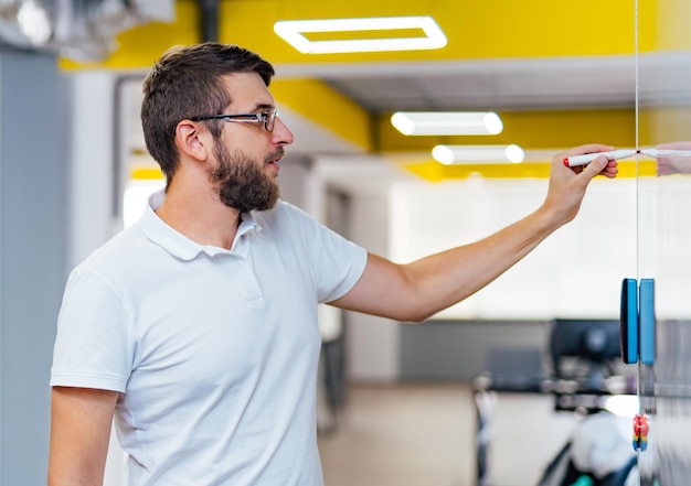 Un hombre barbudo serio con anteojos está dibujando una estrategia de negocios en la pizarra blanca con un marcador Programador de computadoras en una camiseta blanca con un marcador escribiendo en la pizarra en una oficina moderna