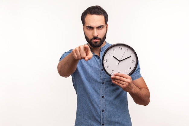 Hombre barbudo serio con camisa azul señalando con el dedo a la cámara sosteniendo un gran reloj de pared en la mano, tiempo de acción, motivación. Disparo de estudio interior aislado sobre fondo blanco.