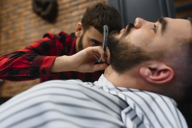 Hombre barbudo serio conseguir corte de pelo de barba con una navaja de afeitar por peluquero mientras está sentado en una silla en la barbería. Tema de barbería