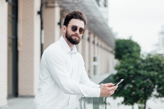 Un hombre barbudo, serio y elegante con camisa blanca y gafas de sol de pie en las calles de la ciudad cerca de la oficina moderna con teléfono móvil