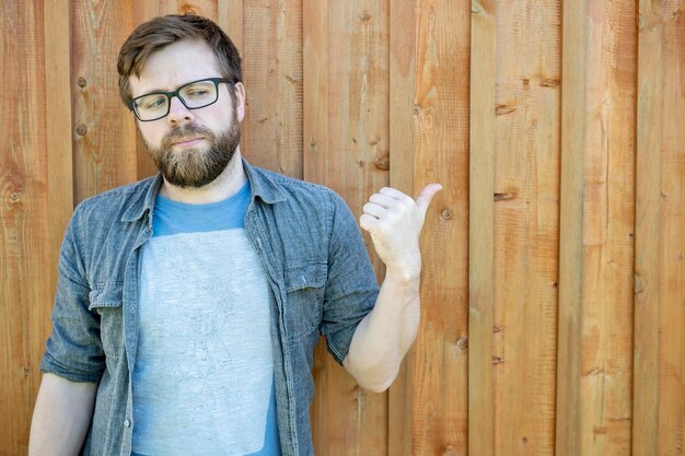 Un hombre barbudo serio con gafas muestra su pulgar en una pared vieja de madera y mira hacia otro lado