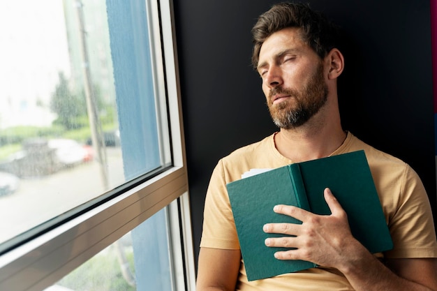 Hombre barbudo sobrecargado de trabajo durmiendo con un libro en la mano sentado cerca de la ventana
