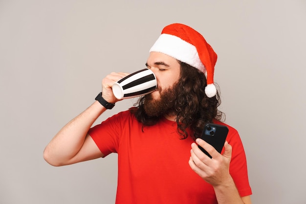 El hombre barbudo con sombrero de Navidad está bebiendo de una taza y sosteniendo su teléfono