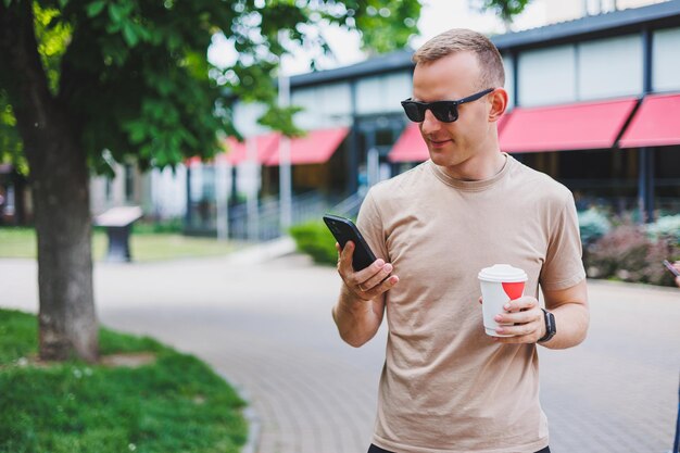 Un hombre barbudo y sonriente con camisa blanca y gafas de sol parado en las calles de la ciudad y llamando por teléfono móvil