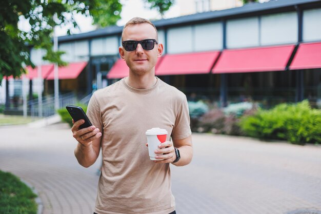 Un hombre barbudo y sonriente con camisa blanca y gafas de sol parado en las calles de la ciudad y llamando por teléfono móvil