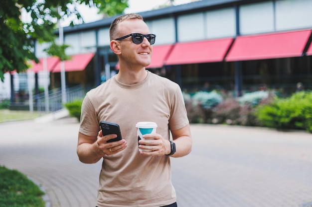 Un hombre barbudo y sonriente con camisa blanca y gafas de sol parado en las calles de la ciudad y llamando por teléfono móvil