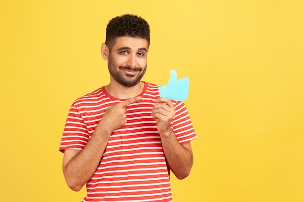 Hombre barbudo sonriente con camiseta a rayas señalando con el dedo los pulgares azules hacia arriba, como un botón de contador, pidiendo calificar las publicaciones en las redes sociales. Disparo de estudio interior aislado sobre fondo amarillo