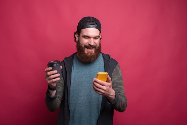 Hombre barbudo sonriente está mirando su teléfono mientras sostiene una taza de papel sobre fondo rosa.