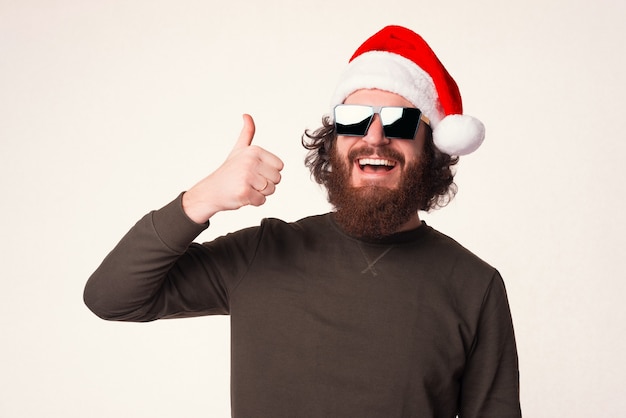 Hombre barbudo sonriente está mostrando el pulgar hacia arriba sobre fondo blanco en Navidad.