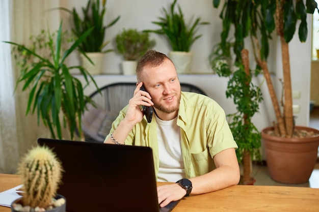 Hombre barbudo sonriente hablando por teléfono usando una computadora portátil