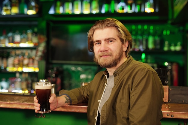 Hombre barbudo sonriente joven en ropa casual mirándote mientras está de pie frente a la cámara contra la barra con bebidas alcohólicas y tomando un vaso de cerveza