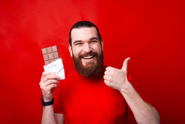 Hombre barbudo sonriente sosteniendo una barra de chocolate y mostrando el pulgar hacia arriba, buenos dulces