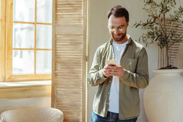 Hombre barbudo sonriente sosteniendo un teléfono inteligente usando una aplicación móvil para comprar en línea en casa