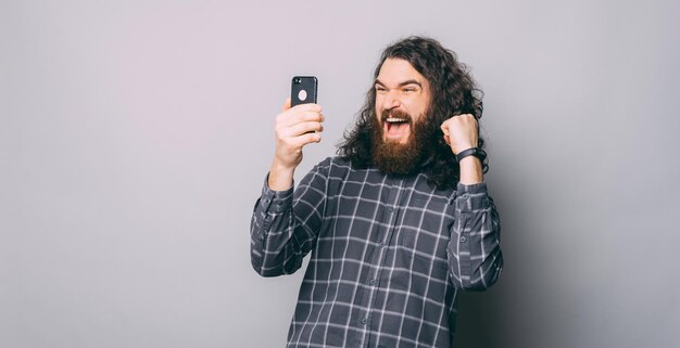 Hombre barbudo sorprendido con cabello largo celebrando y mirando smartphone