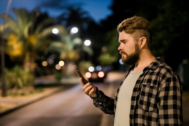 Hombre barbudo sosteniendo mensajes de texto de desplazamiento en su teléfono celular en la calle de noche llama a un taxi en una aplicación