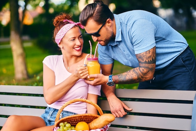 Hombre barbudo tatuado y mujer pelirroja están haciendo un picnic en un banco en un parque.