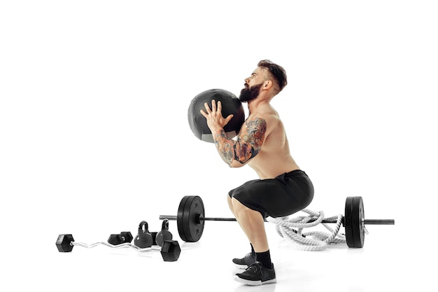 Hombre barbudo tatuado muscular ejerciendo pesas de fitness Balón Medicinal en estudio aislado sobre fondo blanco