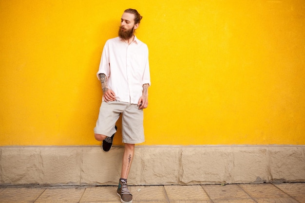 Hombre barbudo tatuado en la pared amarilla posando al aire libre