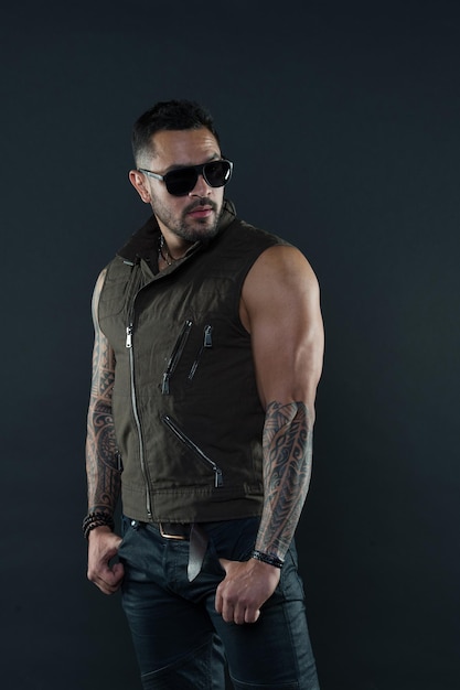 Hombre barbudo con tatuaje en brazos fuertes. Modelo de tatuaje con barba en la cara sin afeitar. Hombre tatuado con bíceps y tríceps. Macho de moda en gafas de sol de moda. Estilo de moda y tendencia.